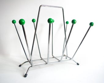 Peut inclure: Un porte-revues chromé avec des boules vertes décoratives. Le porte-revues a une base rectangulaire et une poignée en haut. Le design présente des tiges verticales avec des sphères vertes, créant une esthétique moderne du milieu du siècle. Hauteur d'environ 30 cm.
