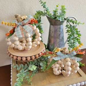 May include: Two wooden bead pumpkins with plaid ribbon and fall foliage accents on a tiered wooden tray with a white bead trim. The tray is on a wooden table with a galvanized metal pitcher and greenery.