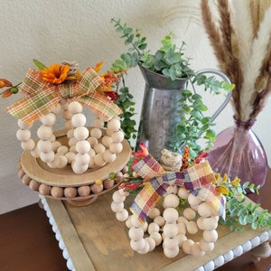 May include: Two wooden bead pumpkins decorated with fall-themed fabric bows. The pumpkins are on wooden stands and are surrounded by greenery.