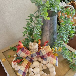 May include: A wooden bead pumpkin with a plaid bow and fall-colored floral accents. The pumpkin is sitting on a wooden surface with a white and gray background.