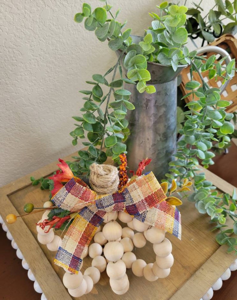 May include: A wooden bead pumpkin with a plaid bow and fall-colored floral accents. The pumpkin is sitting on a wooden surface with a white and gray background.