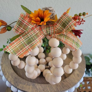 May include: A wooden bead pumpkin with a plaid ribbon and fall-colored flowers. The pumpkin is sitting on a wooden tiered tray.