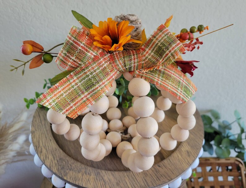 May include: A wooden bead pumpkin with a plaid ribbon and fall-colored flowers. The pumpkin is sitting on a wooden tiered tray.