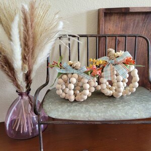May include: Two white wooden bead pumpkins decorated with fall-colored floral accents and blue and white plaid ribbon. The pumpkins are sitting on a metal bench with a light blue cushion.