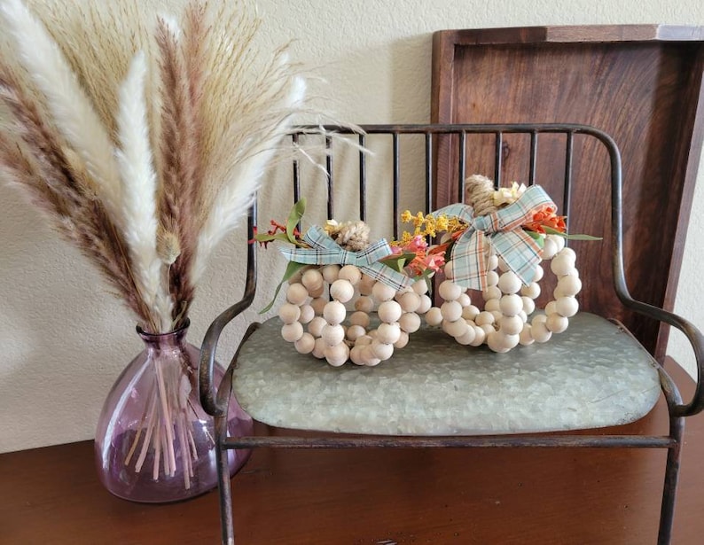 May include: Two white wooden bead pumpkins decorated with fall-colored floral accents and blue and white plaid ribbon. The pumpkins are sitting on a metal bench with a light blue cushion.