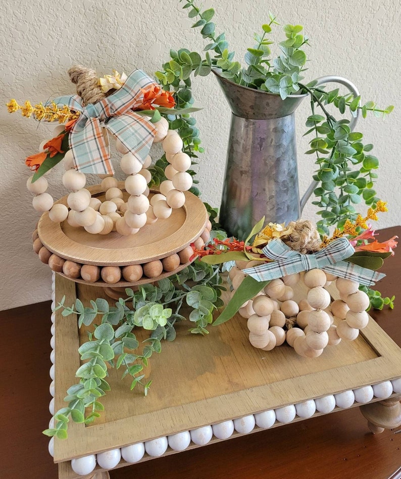 May include: Two wooden bead pumpkins with plaid ribbon and fall foliage accents on a tiered wooden tray with a white bead trim. The tray is on a wooden table with a galvanized metal pitcher and greenery.