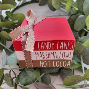 May include: A stack of three wooden blocks with a red, white, and brown color scheme. The blocks are tied together with a brown and white ribbon. The top block reads "CANDY CANES", the middle block reads "MARSHMALLOWS", and the bottom block reads "HOT COCOA".