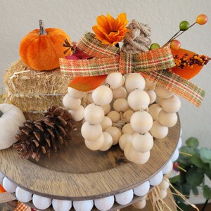 May include: A wooden bead pumpkin with a plaid ribbon and fall-themed flowers. The pumpkin is sitting on a tiered tray with a pine cone and a small orange pumpkin.