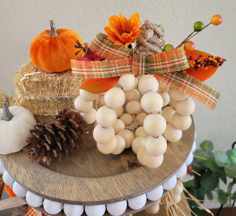 May include: A wooden bead pumpkin with a plaid ribbon and fall-themed flowers. The pumpkin is sitting on a tiered tray with a pine cone and a small orange pumpkin.