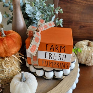 May include: A wooden tiered tray with a farmhouse style sign that reads "Farm Fresh Pumpkins". The tray is decorated with white pumpkins, a brown and orange pumpkin, and greenery.