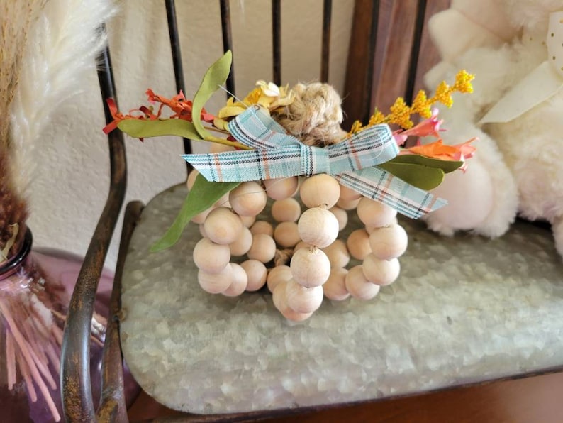 May include: A wooden bead pumpkin with a blue and white plaid bow and fall-colored floral accents.