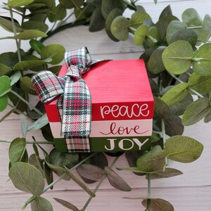 Op de afbeelding: Een stapel van drie houten blokken geverfd in rood, wit en groen. De blokken zijn versierd met de woorden "peace", "love" en "JOY" in witte letters. De blokken zijn aan elkaar gebonden met een rood-groen geruit lint.
