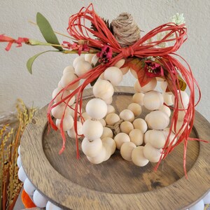 May include: A wooden pumpkin decoration made of round beads, with a red and white bow and dried flowers. The pumpkin is sitting on a wooden base.