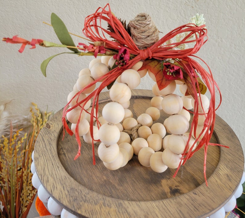 May include: A wooden pumpkin decoration made of round beads, with a red and white bow and dried flowers. The pumpkin is sitting on a wooden base.