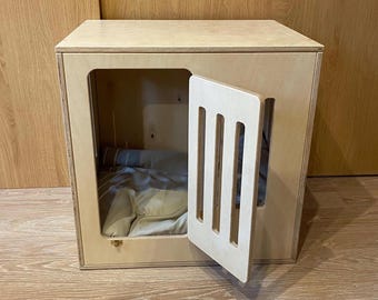 Può includere: Una cassa per animali domestici in legno chiaro con un design rettangolare. La cassa presenta una porta con ritagli verticali e un letto all'interno. La porta è parzialmente aperta, rivelando l'interno. La cassa è in legno chiaro.