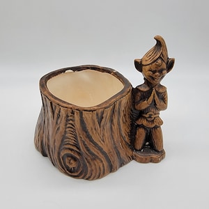 May include: A ceramic planter shaped like a tree stump with a small elf figurine attached. The planter is brown and has a textured surface. The elf is wearing a brown hat and a brown tunic.