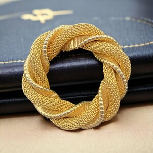 May include: A circular brooch featuring a braided design with a textured gold-tone finish. The piece is composed of interwoven mesh-like strands that create a continuous, knotted loop, showcasing a metallic luster and intricate metalwork details.