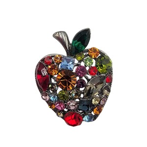 May include: A silver-toned apple-shaped brooch encrusted with a variety of colorful gemstones. The brooch features a green leaf and stem, with the apple body filled with red, orange, yellow, blue, and purple stones.