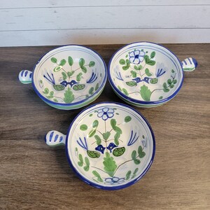 May include: Three ceramic bowls with handles, featuring a hand-painted floral design in blue and green on a white background. Each bowl has a blue rim and a small handle. The bowls are arranged on a wooden surface.