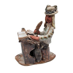 Peut inclure: Une figurine en céramique d'une femme portant un chapeau et assise à un bureau avec une plume et des livres. La figurine est peinte dans des tons de brun, blanc et vert.