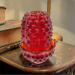 Pode incluir: Um castiçal de vidro vermelho com um design de pregos. O castiçal tem uma base alargada e um corpo cilíndrico. O vidro é de um vermelho profundo e transparente. Está sobre uma mesa de madeira.