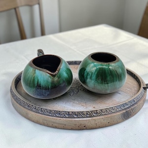 Puede incluir: Un juego de azucarero y jarra de cerámica verde y marrón. La jarra tiene un pico y un asa. El juego se encuentra sobre una bandeja redonda de madera con un borde decorativo. El interior de la jarra y el cuenco es negro.