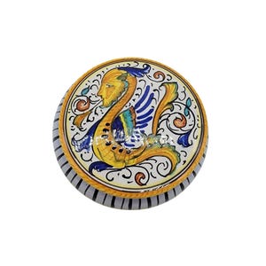 May include: A round ceramic trinket box with a hand-painted design. The lid features a mythical creature with a yellow head, blue wings, and a yellow tail. The box has a white background with decorative swirls and a striped edge.