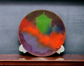Puede incluir: Un plato redondo de cerámica con un diseño abstracto colorido. El plato está pintado en tonos de naranja, verde, azul y morado. El plato está en un estante de madera.