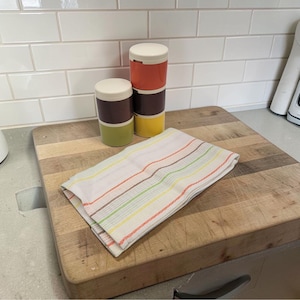 May include: A stack of three colorful food storage containers with white lids. The containers are orange, brown, and yellow. A folded striped kitchen towel rests on a wooden cutting board.