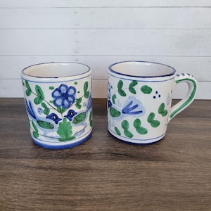 May include: Two ceramic mugs with a white base, adorned with blue floral and green leaf designs. One mug has a handle with a green accent. The rims and bases are painted blue. The mugs are on a dark wooden surface.