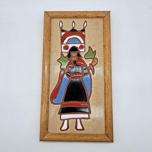 Puede incluir: Un azulejo de cerámica enmarcado con una representación colorida de una mujer nativa americana con una corona y sosteniendo una planta verde. El azulejo está sobre un fondo beige.