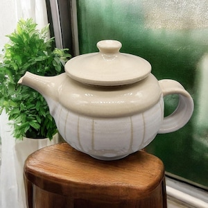 Può includere: Una teiera in ceramica bianca con un coperchio e un manico. La teiera ha un design testurizzato e a coste e si trova su una superficie di legno.