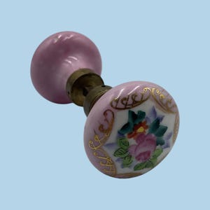 May include: A round porcelain door knob featuring a light pink base color with a hand-painted floral motif in shades of pink, green, and blue, accented by gold filigree detailing. The knob is attached to a central brass-toned metal mounting post.
