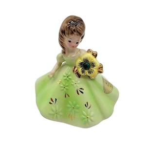 May include: A porcelain figurine of a girl wearing a green dress with a yellow flower featuring a green gem in the centre. The girl is holding the flower in her right hand. The dress is decorated with gold trim and small green flowers.