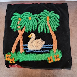 May include: Black cushion with a tufted design depicting a swan on a lake, flanked by palm trees. The palm trees have green fronds and brown trunks. The swan is cream-coloured with a yellow beak. The lake is blue.