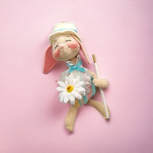 May include: A plush bunny doll wearing a white hat and a blue and white dress, holding a white flower and a white stick. The doll is on a pink background.