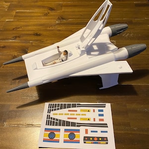 Könnte beinhalten: Ein weißes Plastikmodell eines Raumschiffs mit einem grauen Cockpit und einer Figur im Inneren. Das Raumschiff hat einen angehobenen Flügel und zwei graue Triebwerke. Ein Bogen Aufkleber mit schwarzen, weißen, roten, gelben und blauen Designs ist enthalten.