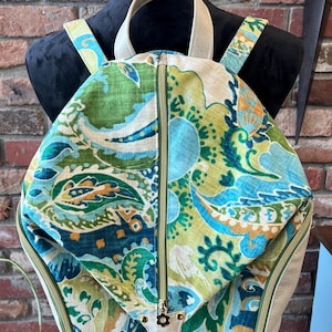 May include: A colorful backpack with a floral pattern in shades of green, blue, and beige. The backpack has a zippered closure, a top handle, and adjustable shoulder straps. The bag is displayed on a black mannequin.