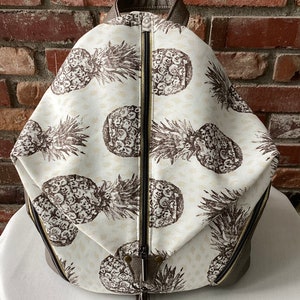 May include: A white canvas backpack with a brown leather trim and a zipper closure. The backpack has a pineapple print pattern.