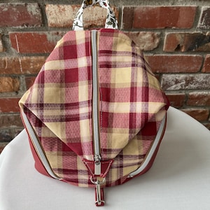 May include: A red and beige plaid backpack with a floral patterned handle. The backpack has two zipper closures and a silver zipper pull.