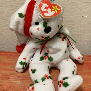 Peut inclure: Ours en peluche blanc Beanie Baby portant un bonnet de Père Noël rouge et blanc, avec un motif de houx vert et rouge. L'ours a des yeux noirs en forme de boutons et un nez rouge. L'ours est assis sur une surface en bois. L'ours a une étiquette Ty attachée à son oreille qui dit "Beanie Babies Baby".