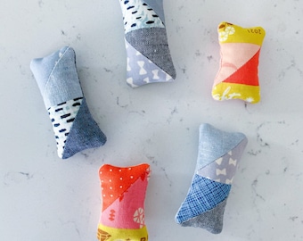 Può includere: Cinque piccoli cuscini rettangolari realizzati in tessuto patchwork. I cuscini sono di vari colori e motivi, tra cui blu, giallo, rosa e rosso. I cuscini sono disposti a semicerchio su una superficie bianca.
