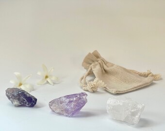 Könnte beinhalten: Drei Kristalle, ein violetter Amethyst, ein klarer Quarz und ein dunkelvioletter Amethyst, liegen auf einer weißen Oberfläche. Im Hintergrund befindet sich ein kleiner, beiger Beutel mit Kordelzug.