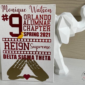 Puede incluir: Un lienzo blanco con texto granate que dice "Monique Watson #9 Orlando Alumnae Chapter Spring 2021 REIGN Supreme Delta Sigma Theta". Un gr&aacute;fico de pato amarillo est&aacute; encima de la palabra "REIGN".