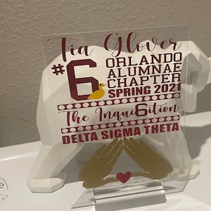 Puede incluir: Placa acr&iacute;lica transparente con texto granate y dorado. La placa incluye el nombre "Tia Glover", "Orlando Alumnae Chapter Spring 2021" y "Delta Sigma Theta". Tambi&eacute;n se muestra un pato dorado y s&iacute;mbolos de manos.
