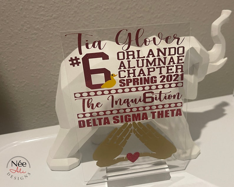 Puede incluir: Placa acr&iacute;lica transparente con texto granate y dorado. La placa incluye el nombre "Tia Glover", "Orlando Alumnae Chapter Spring 2021" y "Delta Sigma Theta". Tambi&eacute;n se muestra un pato dorado y s&iacute;mbolos de manos.