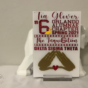 Puede incluir: Placa acr&iacute;lica blanca con texto rojo brillante que dice "Tia Glover #6 Orlando Alumnae Chapter Spring 2021 The Inquisition Delta Sigma Theta." Presenta un pato amarillo y manos doradas con un coraz&oacute;n rojo.