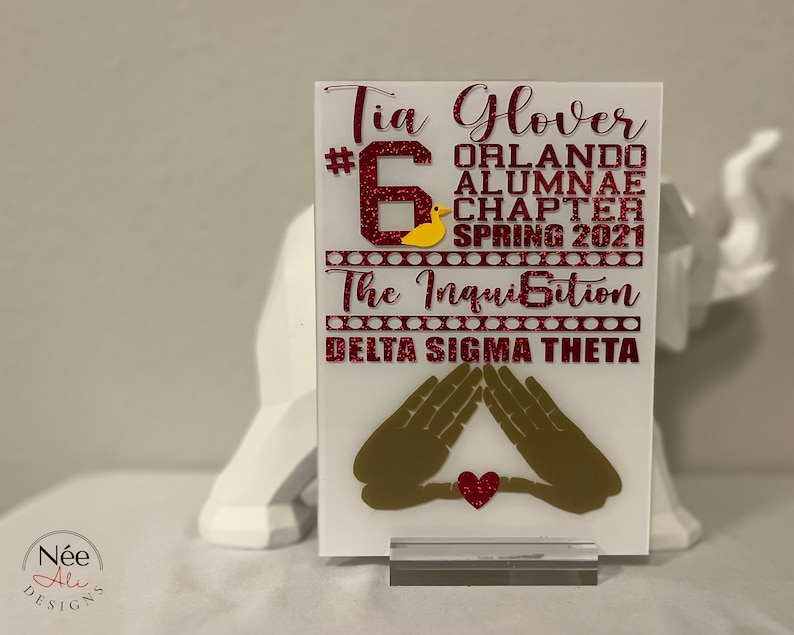 Puede incluir: Placa acr&iacute;lica blanca con texto rojo brillante que dice "Tia Glover #6 Orlando Alumnae Chapter Spring 2021 The Inquisition Delta Sigma Theta." Presenta un pato amarillo y manos doradas con un coraz&oacute;n rojo.