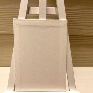 Puede incluir: Un lienzo en blanco, blanquecino, estirado sobre un marco de madera, expuesto en un peque&ntilde;o caballete blanco. El lienzo es rectangular, con costuras visibles en los bordes. El caballete tambi&eacute;n es blanco, con un dise&ntilde;o simple en forma de A.