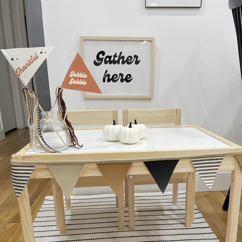 May include: A Thanksgiving-themed table setting with a white tabletop, wooden chairs, and a banner. The table is decorated with white pumpkins and pennant flags that read "Thankful" and "Gobble Gobble." A framed sign says "Gather here."